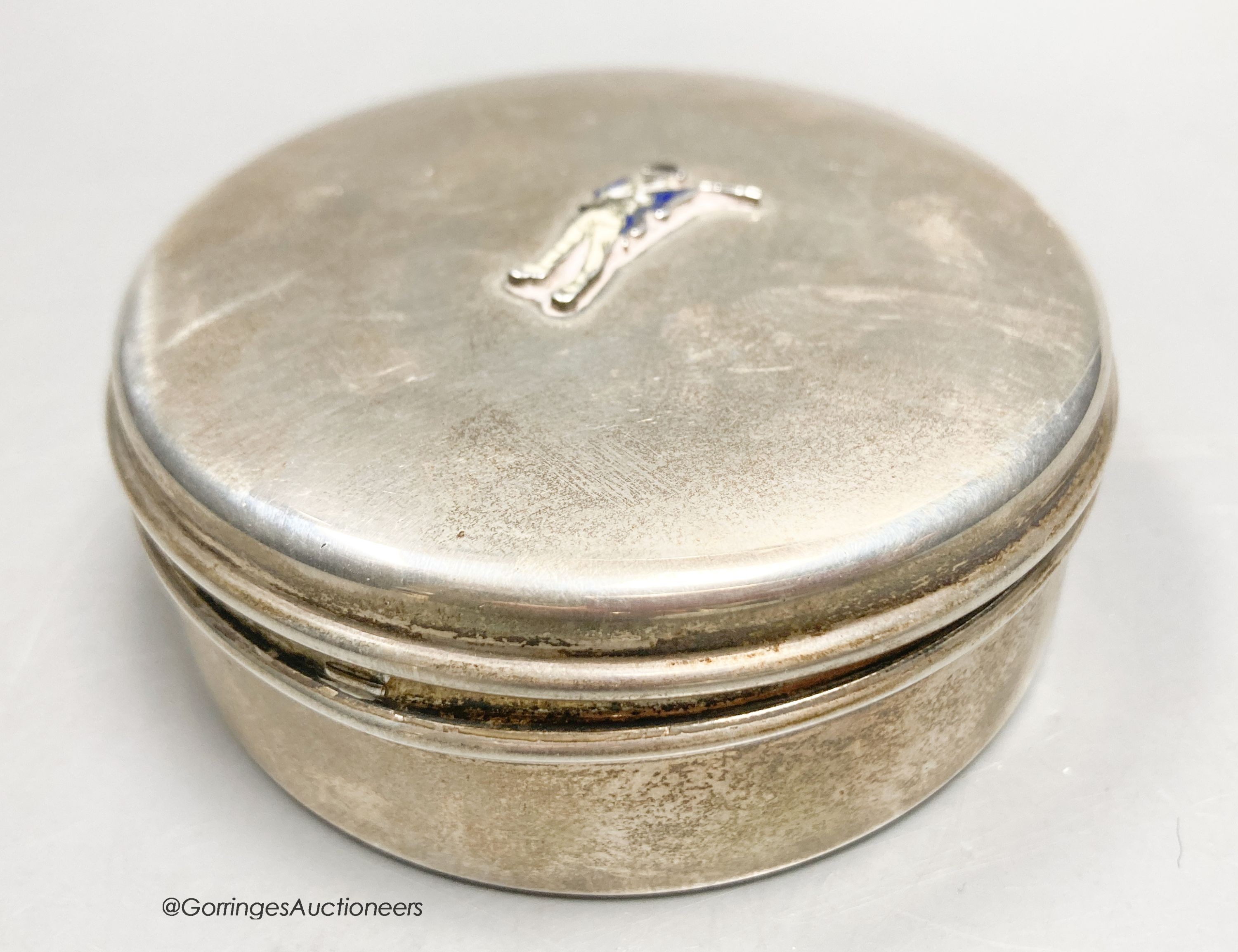 A white metal circular box and hinged cover, with enamelled applique modelled as a soldier, stamped sterling and bearing the stamp 'Cartier', diameter 73mm, gross weight 98 grams.